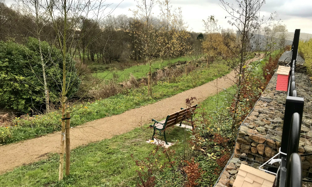 a wooden bench overlooks a path, greenery and canal