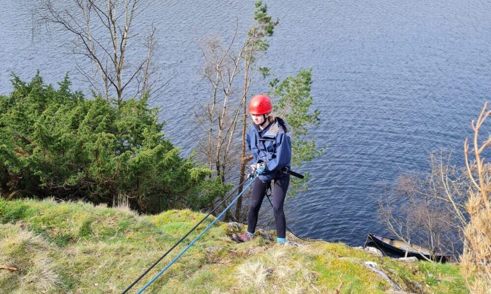 A girl in a red helmet abseils over a lake